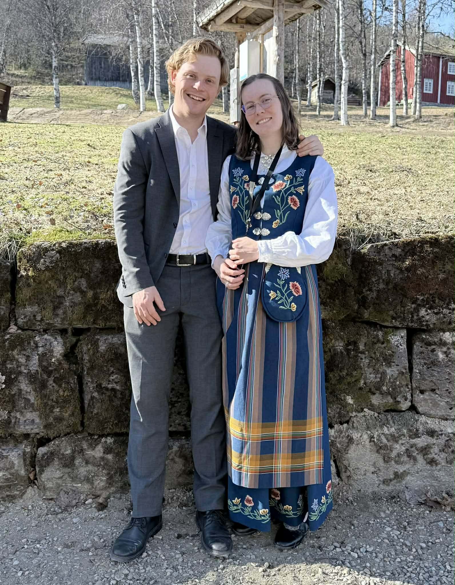 Ola og Kristine i dress og nordlandsbunad foran et rødt stabbur