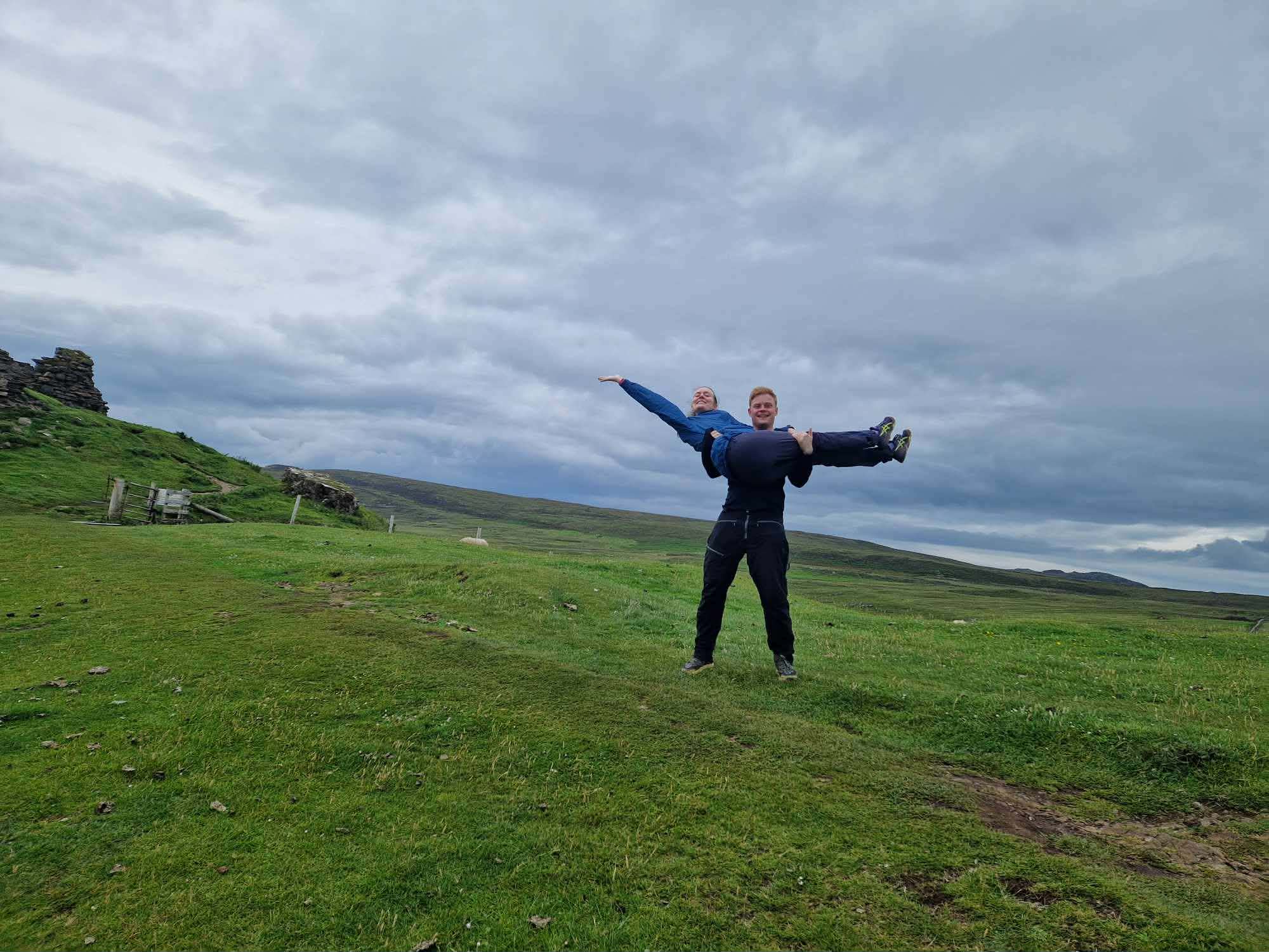 Ola løfter Kristine på en grønn høyde under en dramatisk himmel på Skye
