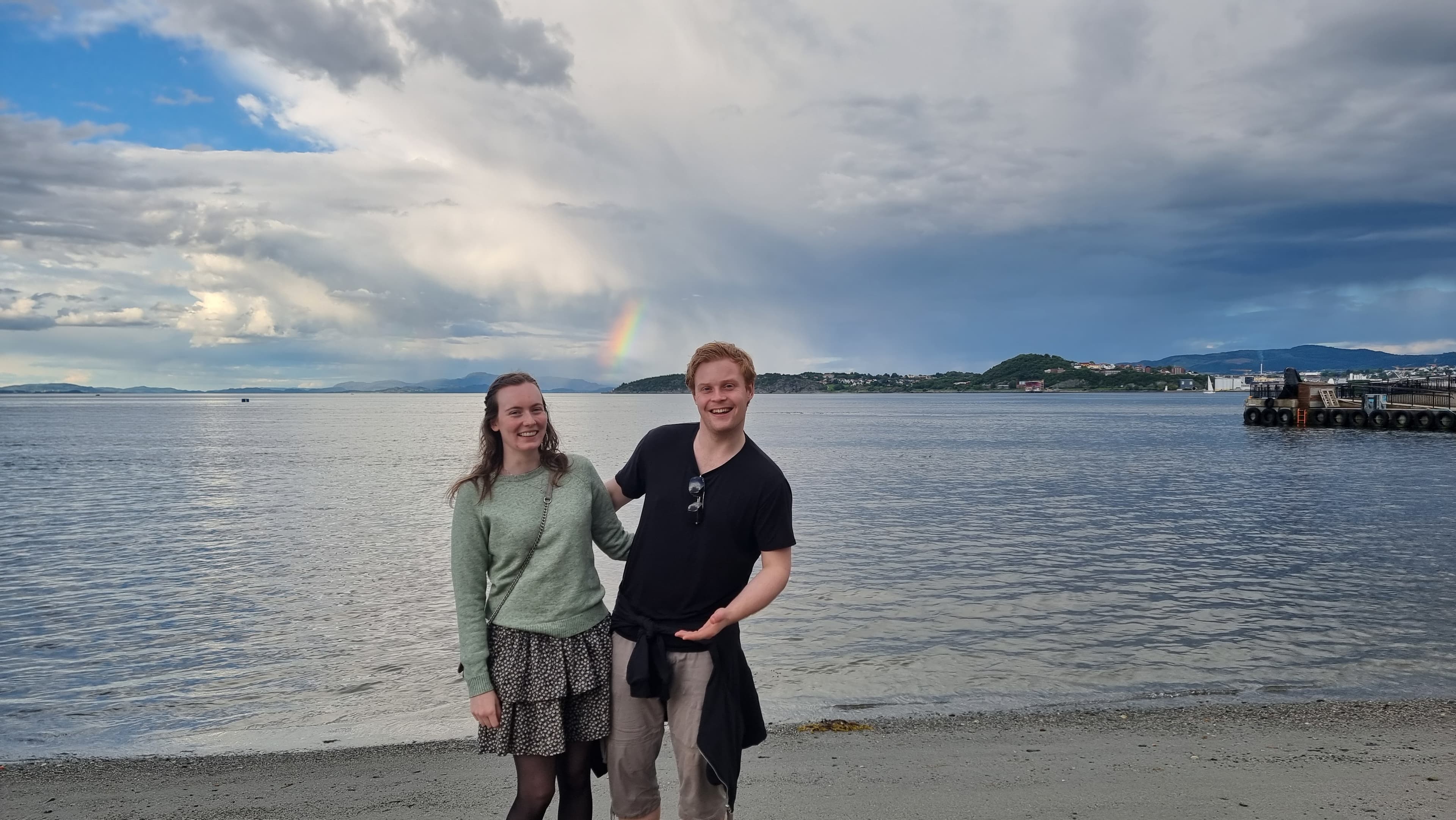 Ola og Kristine står sammen på en strand med sjøen og en regnbue i bakgrunnen